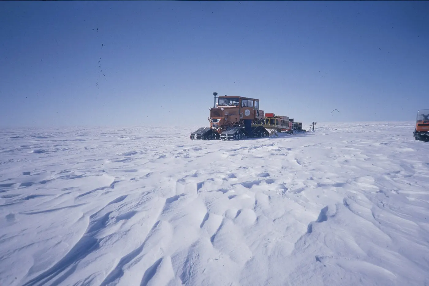 A flat white snowfield stretching into the distance, rippled by the wind, with a snow vehicle parked far off