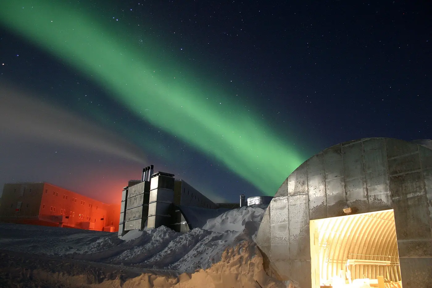 A green aurora stretching like a ribbon across the night sky above snow-buried buildings at the South Pole station