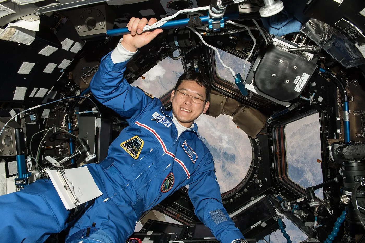 A smiling astronaut in blue floating in front of a large window with Earth visible outside