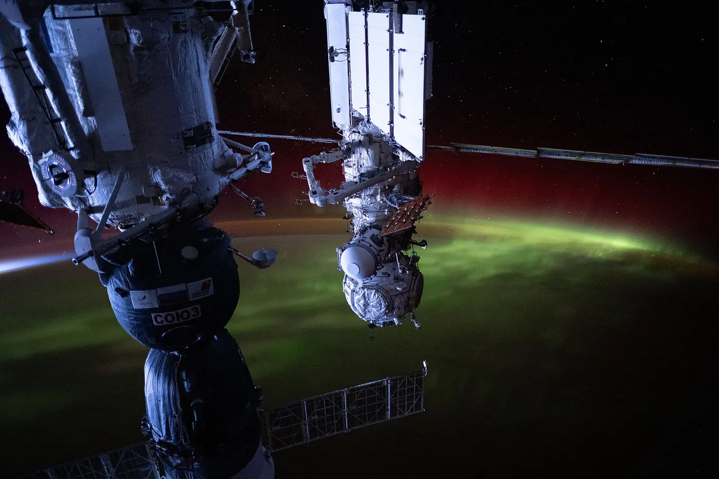 Beyond the body of the space station and a Soyuz spacecraft, green light covers the night side of Earth, with a red glow above it