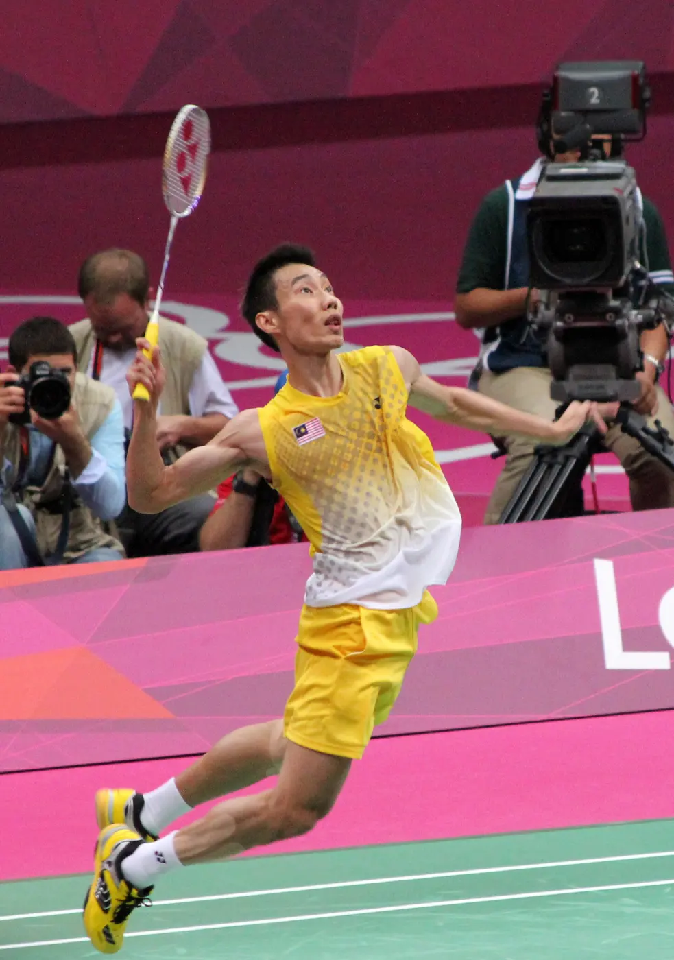 A player in a yellow kit leaps into the air with his racket drawn back over his head, with photographers behind him