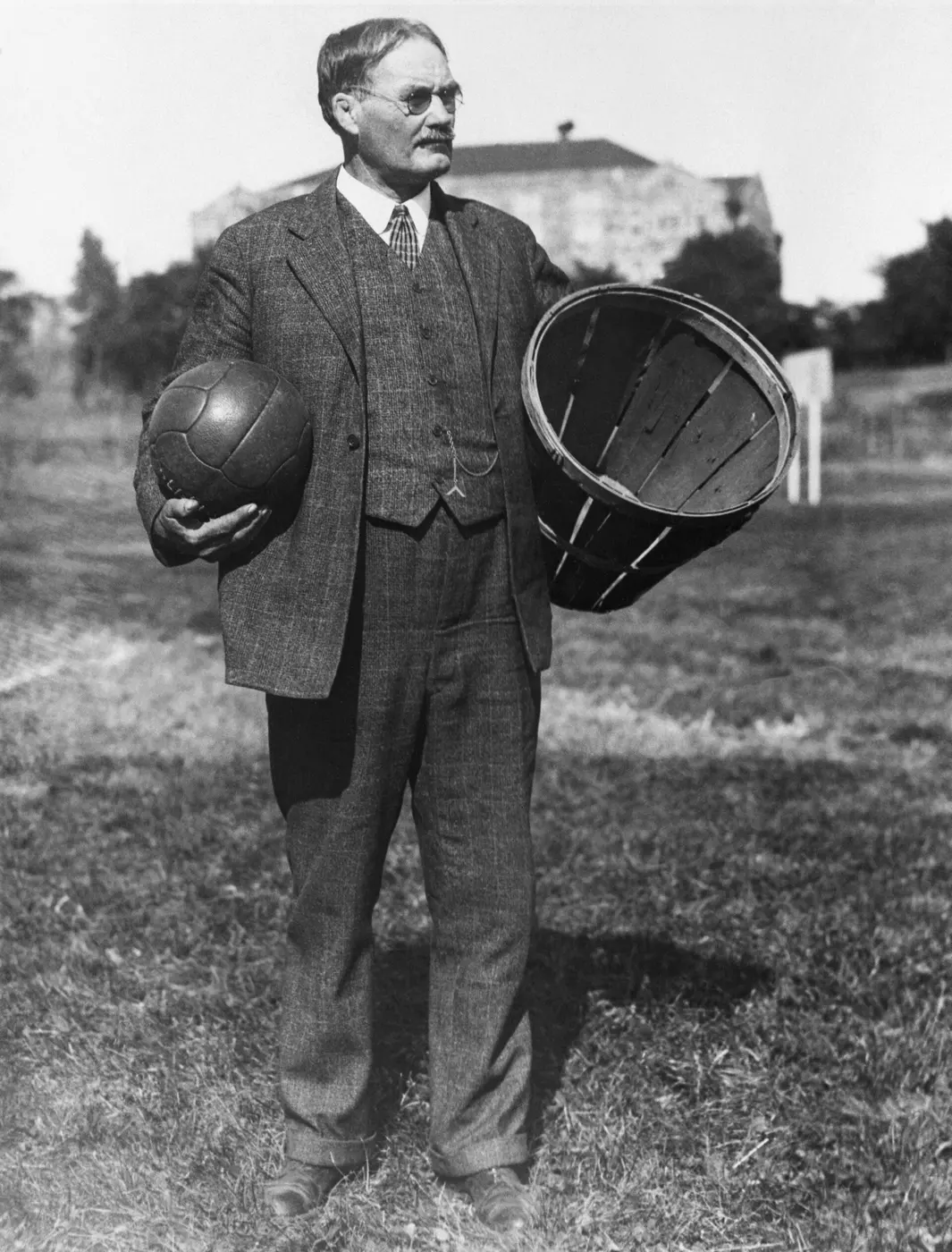 James Naismith in a suit, standing outdoors with a ball in one hand and a wooden basket in the other