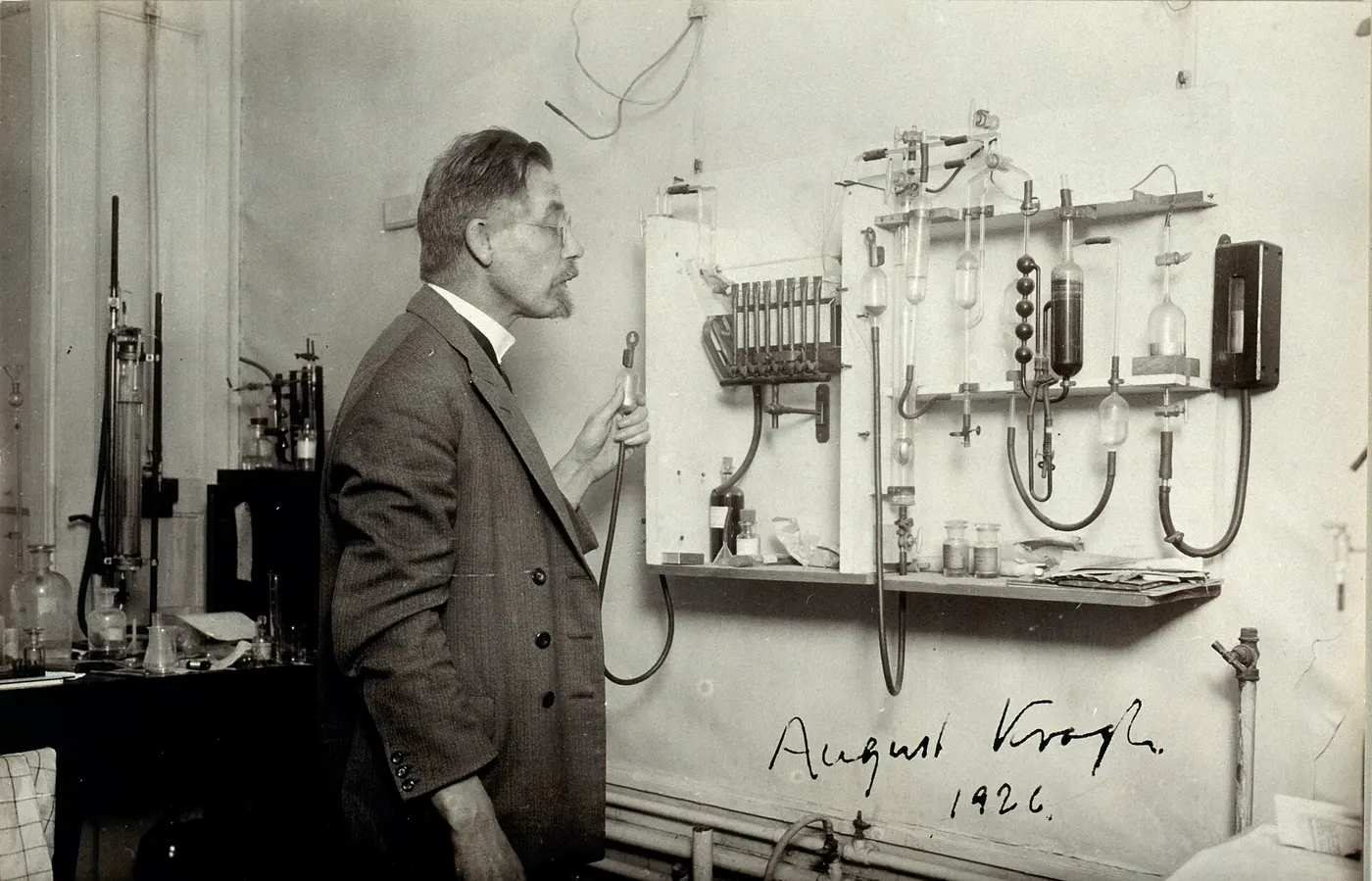 Old photograph of a man in a suit standing in front of a wall lined with laboratory equipment