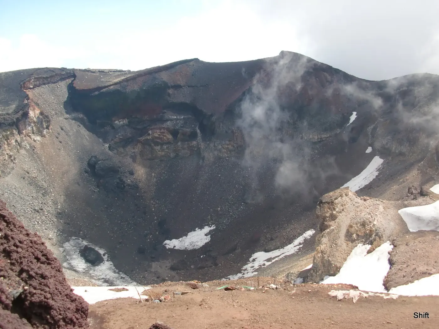 Looking down into the summit crater of Mount Fuji, with snow lingering on rocky slopes and the crater rim stretching away