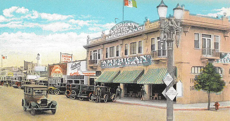 A hand-colored postcard of a street lined with vintage cars, two-story hotels and bars, with a sign reading “CAESAR” in the distance