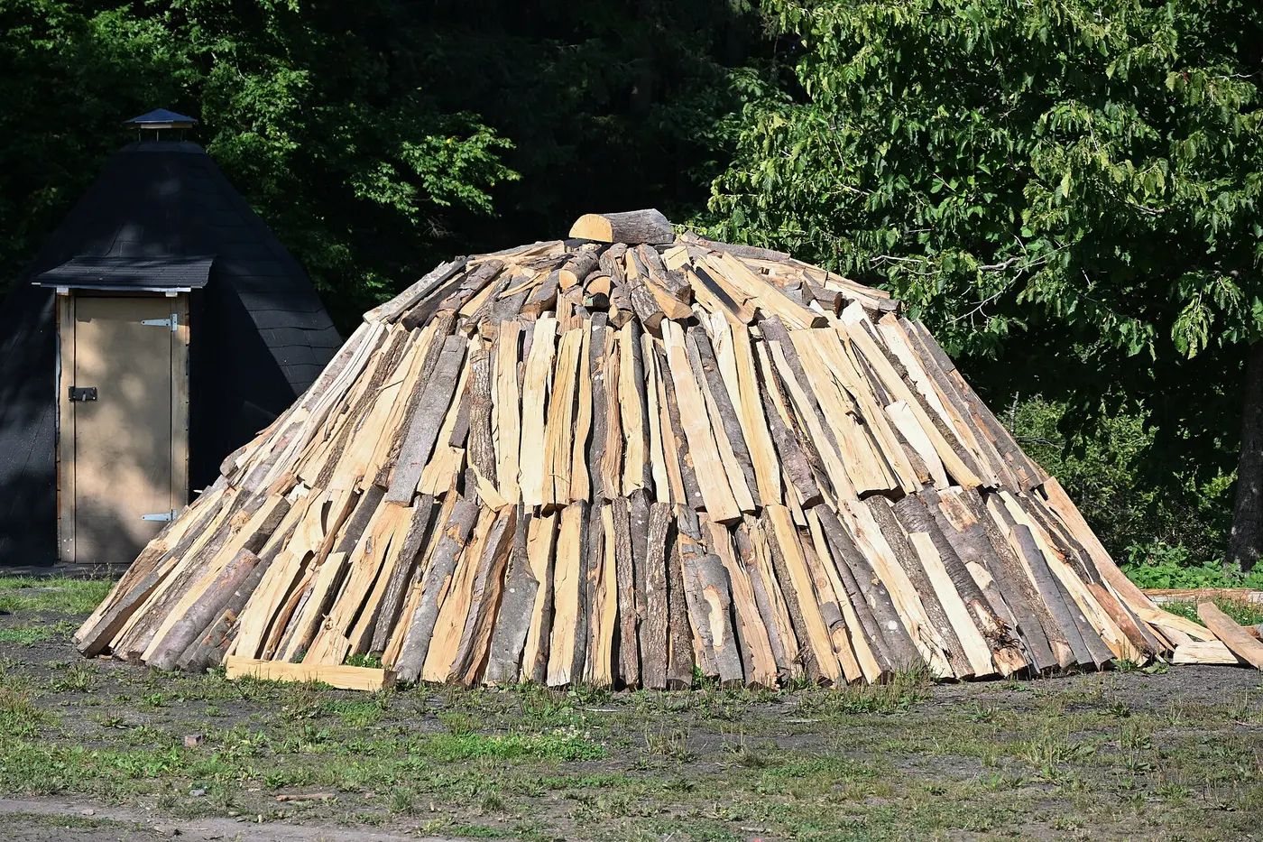 A cone-shaped mound of split logs stacked tightly together in a clearing in front of a forest, ready for charcoal making