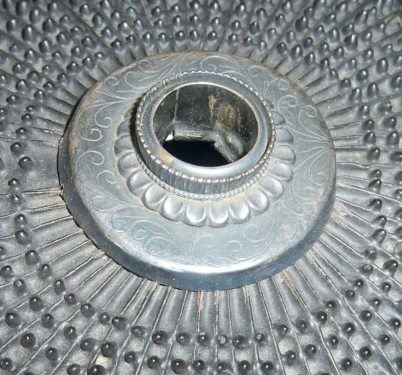 A round opening framed by an ornamental metal fitting at the top of a helmet bowl studded with radiating rivets