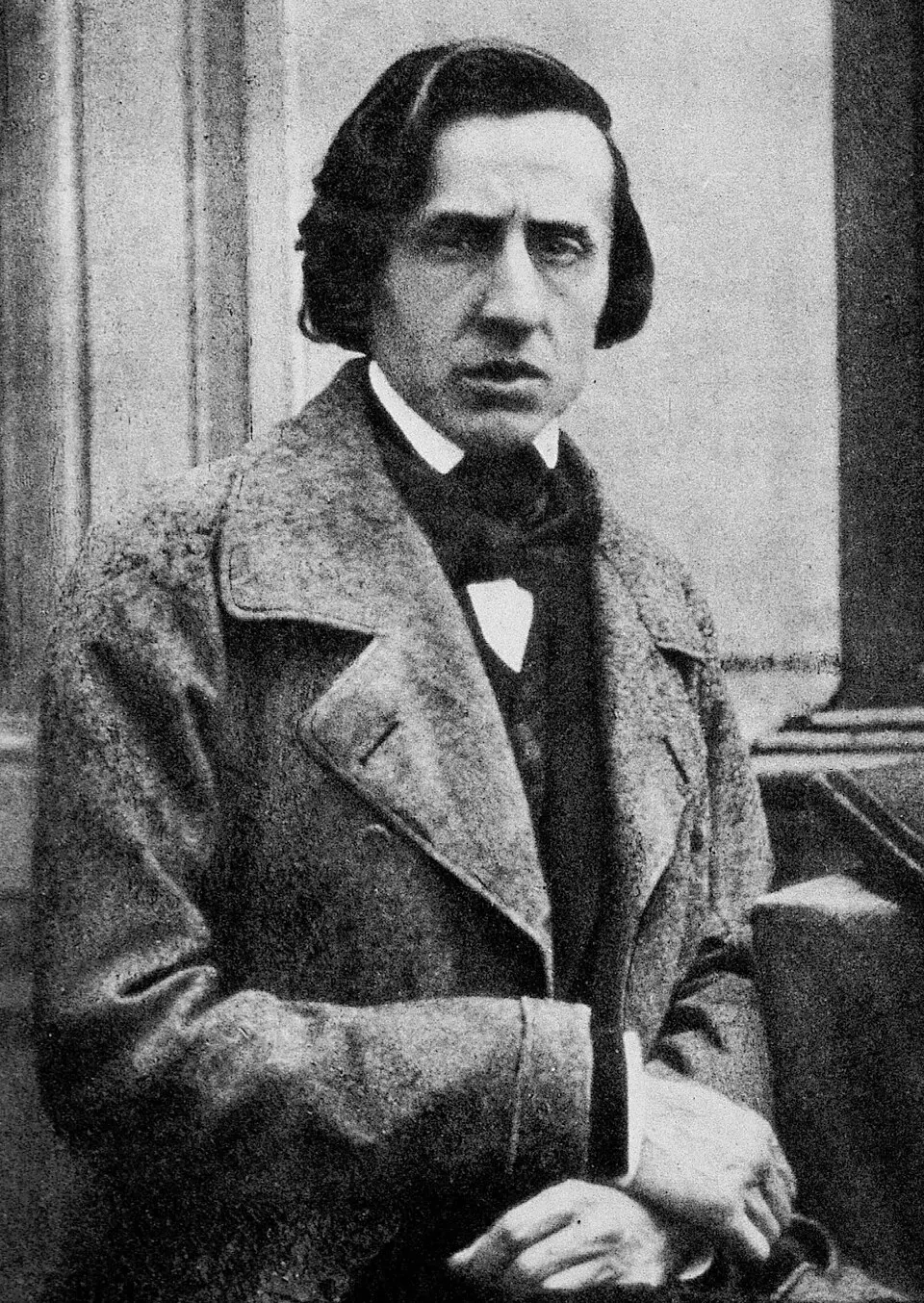 Black-and-white photo of a man with parted, shoulder-length hair in a heavy coat, looking at the camera with a worn expression