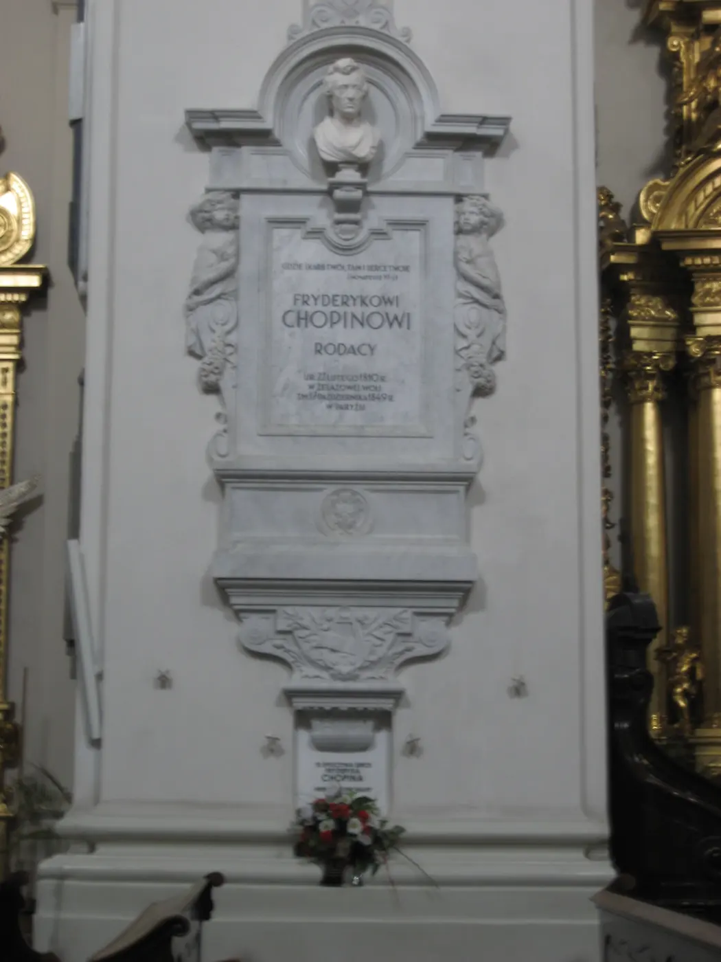 A marble memorial on a white church pillar, with a bust of Chopin at the top and flowers laid below