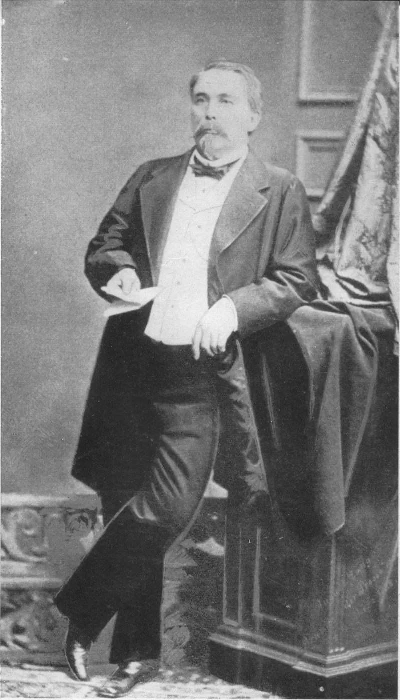 A black-and-white photograph of a mustached man in a frock coat, standing with one elbow resting on a pedestal