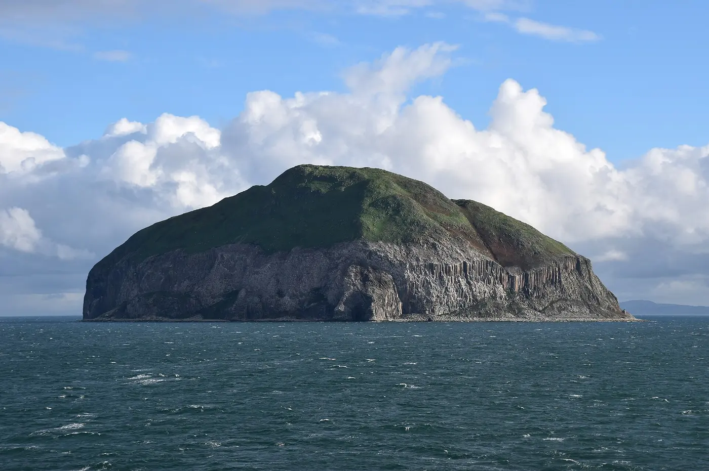 A rocky island shaped like an upturned bowl sits in the sea under a blue sky and white clouds, its cliffs marked with tall columns of rock