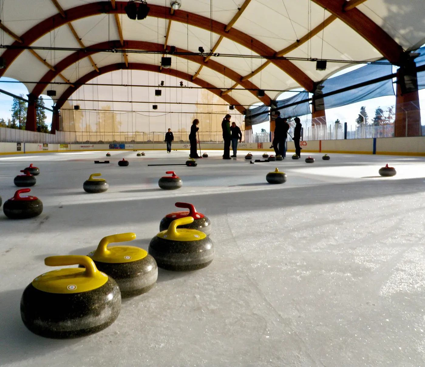 Dark round stones with yellow and red handles scattered on the ice of a covered rink, with people holding brooms in the background