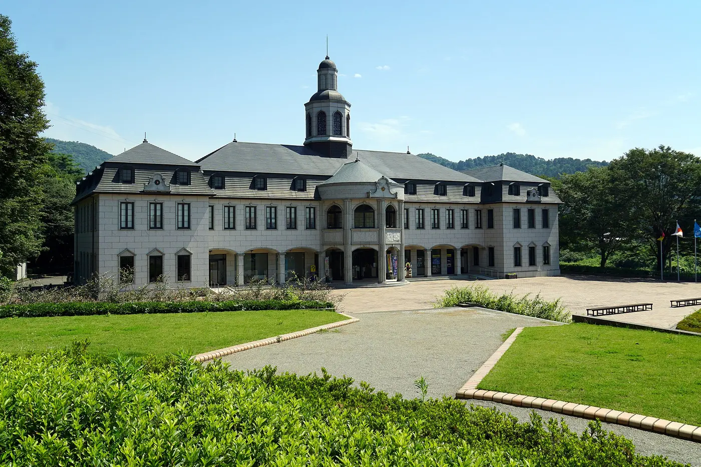 A European-style building with gray roofs, white walls and a central tower, set against green hills with a lawn in front
