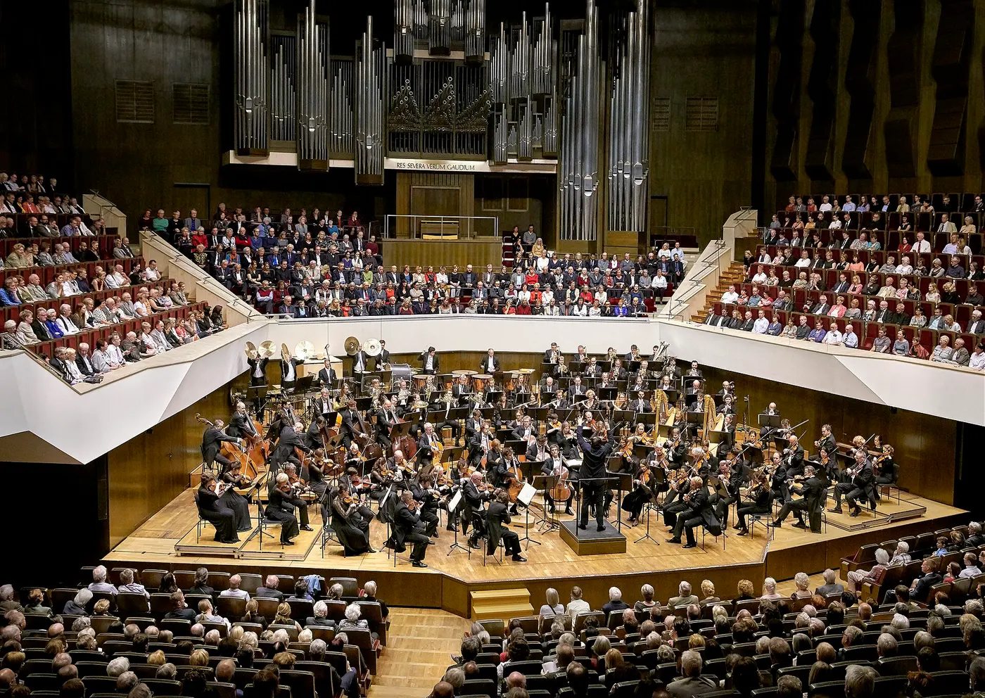 An orchestra performing on a stage in front of a large pipe organ, surrounded by tiers of audience seating