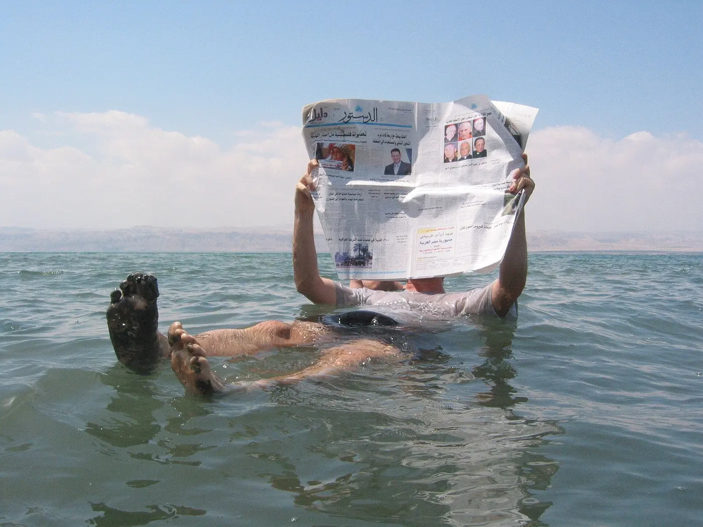 A person floating on their back in the Dead Sea, feet poking out of the water, reading an open newspaper