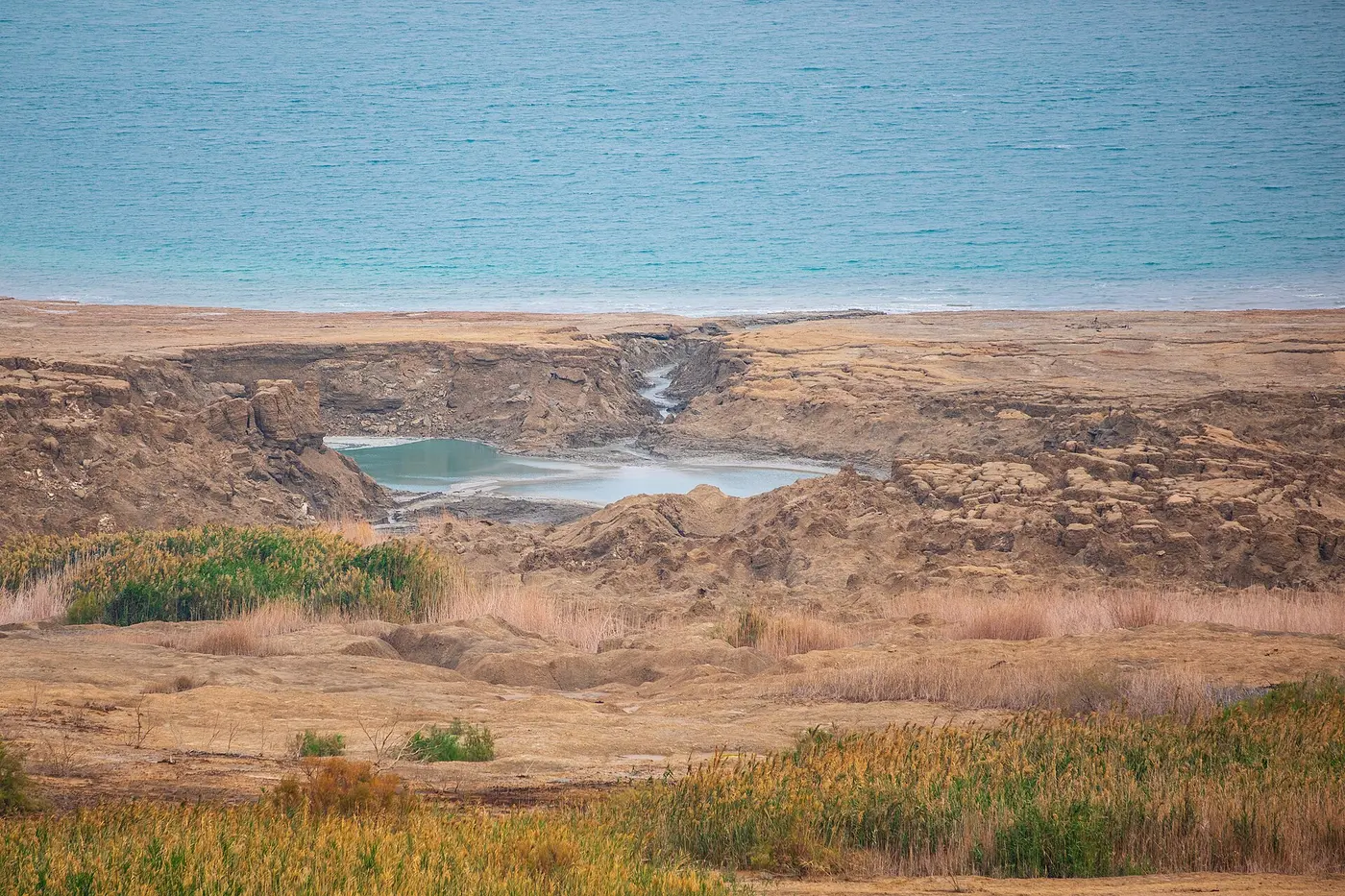 A large collapsed pit with water pooled at the bottom on the shore of the Dead Sea, with the lake’s blue water beyond
