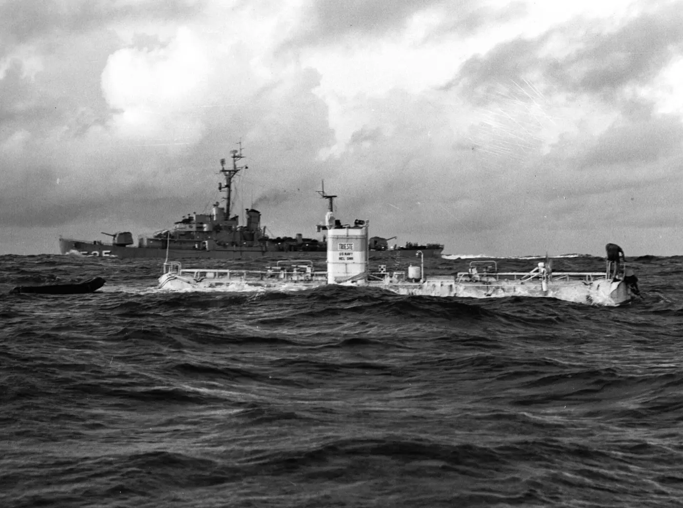 荒れた海面に浮かぶ潜水艇トリエステ号と、背後を進む軍艦の白黒写真