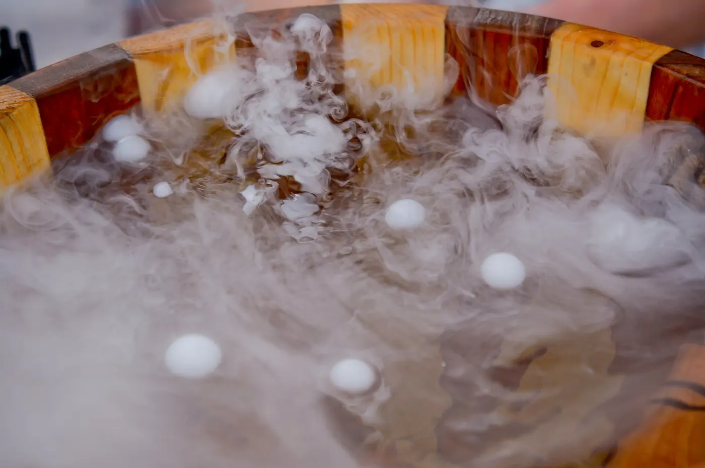 White pellets of dry ice bubble in a wooden bowl of water as clouds of white smoke rise from the surface