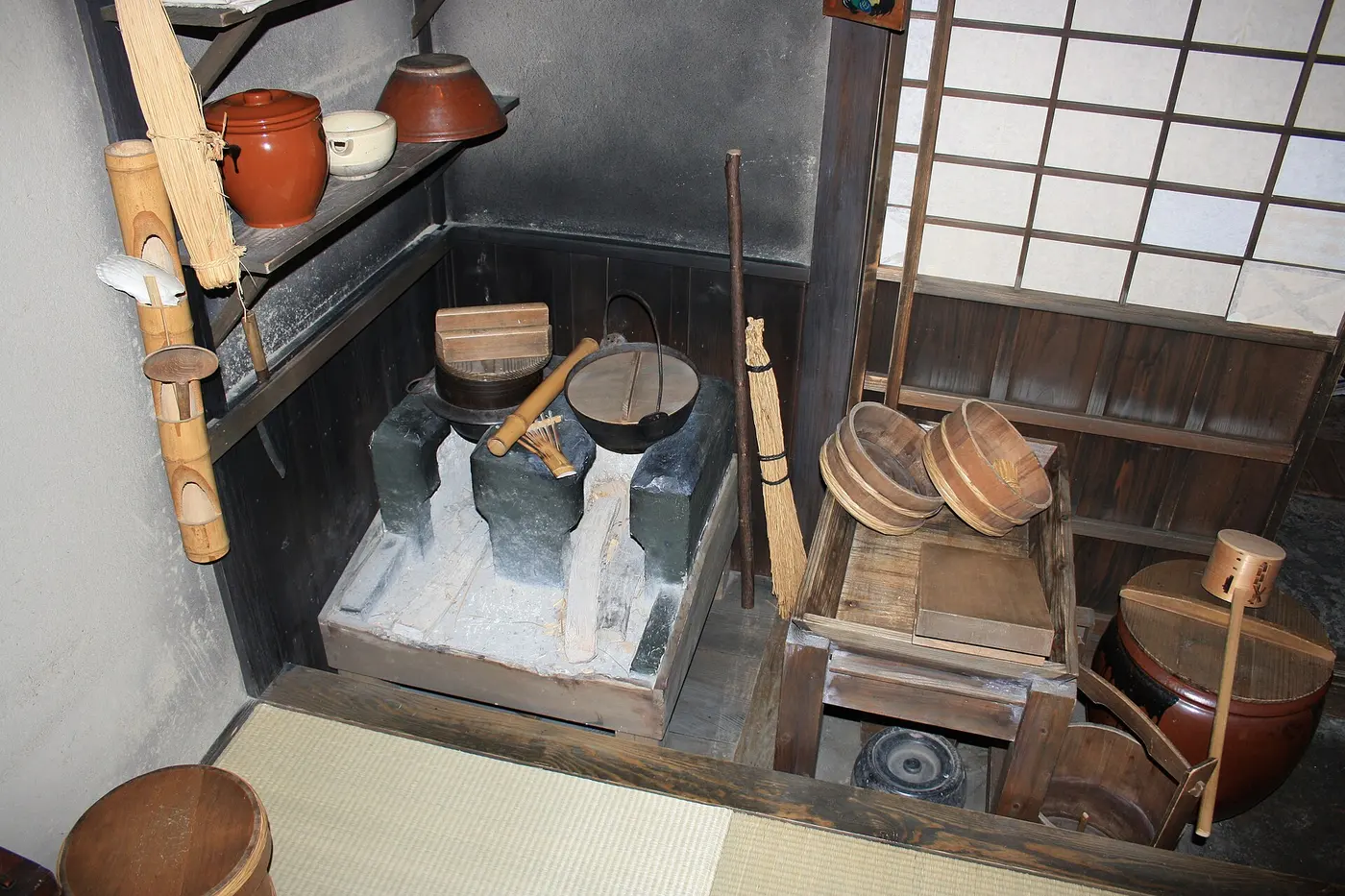 A museum reconstruction of an Edo-period commoner’s kitchen, with a cooking stove, a water jar, and wooden buckets on an earthen floor
