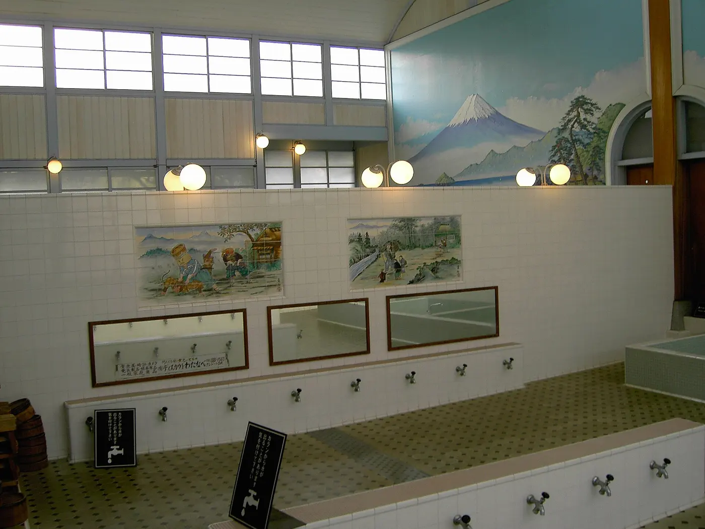 An empty sentō bathing room with a row of taps along the washing area, a tub, and a painting of Mount Fuji on the wall