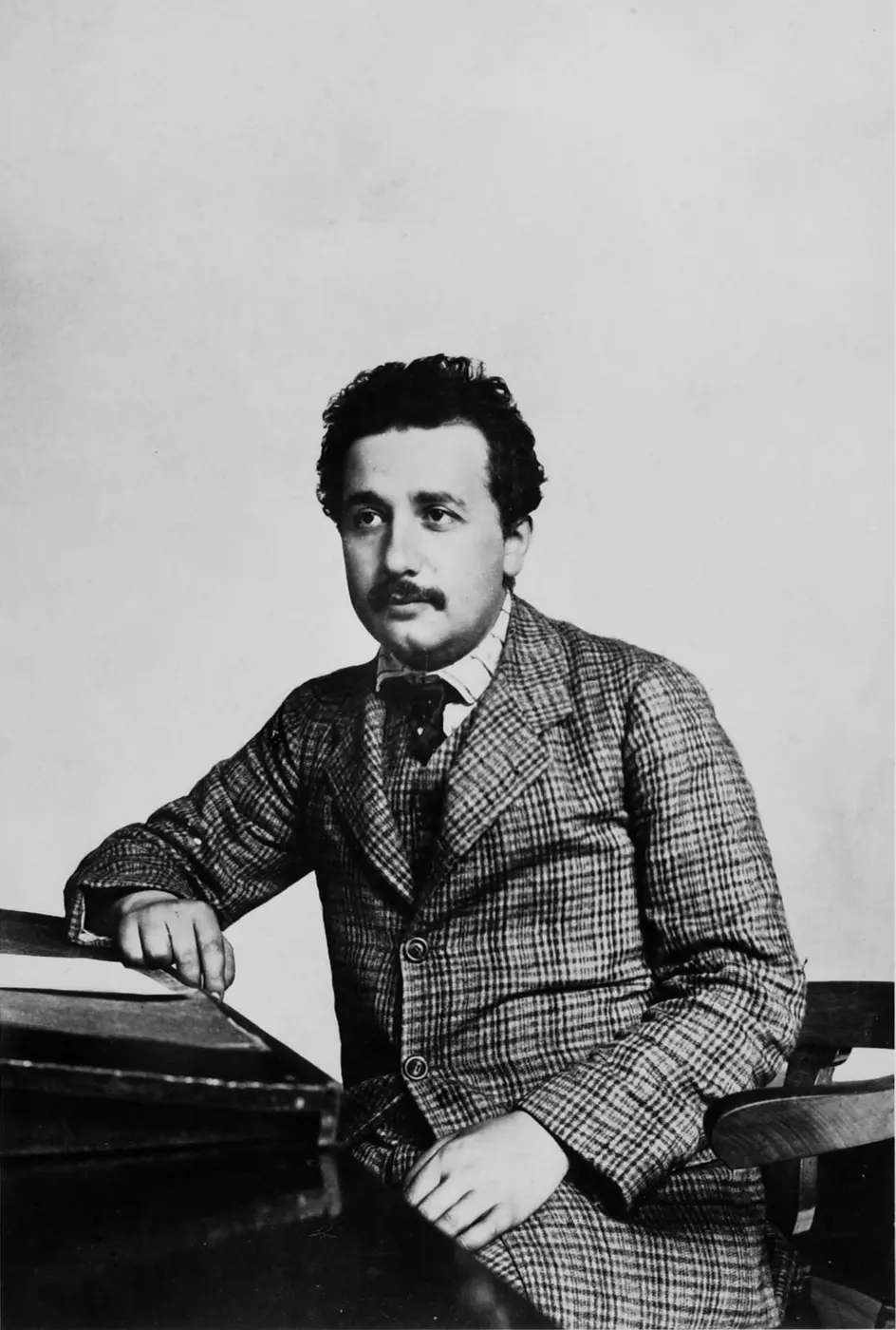 Black-and-white photo of a young man with a mustache in a checked suit, seated at a desk
