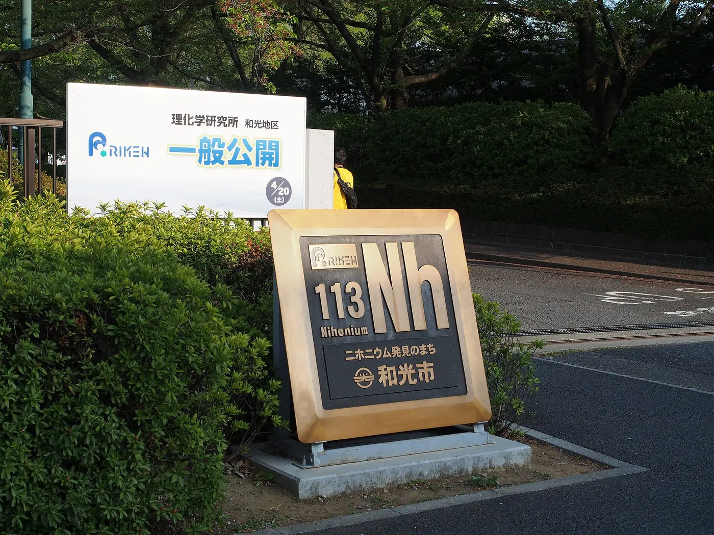 A gold monument in front of a hedge, reading “113 Nh Nihonium,” with a sign saying Wako is the town where nihonium was discovered