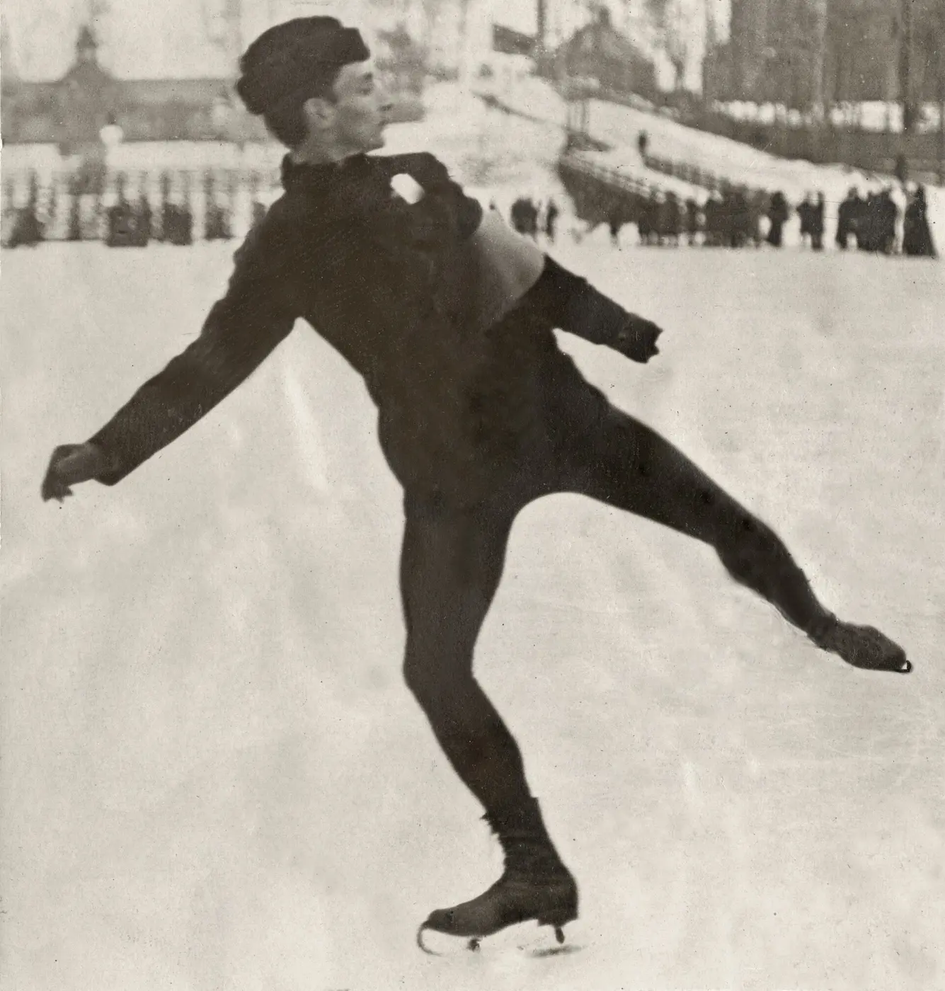 A black-and-white photo of a male skater gliding on one foot on an outdoor rink, arms and one leg stretched out wide