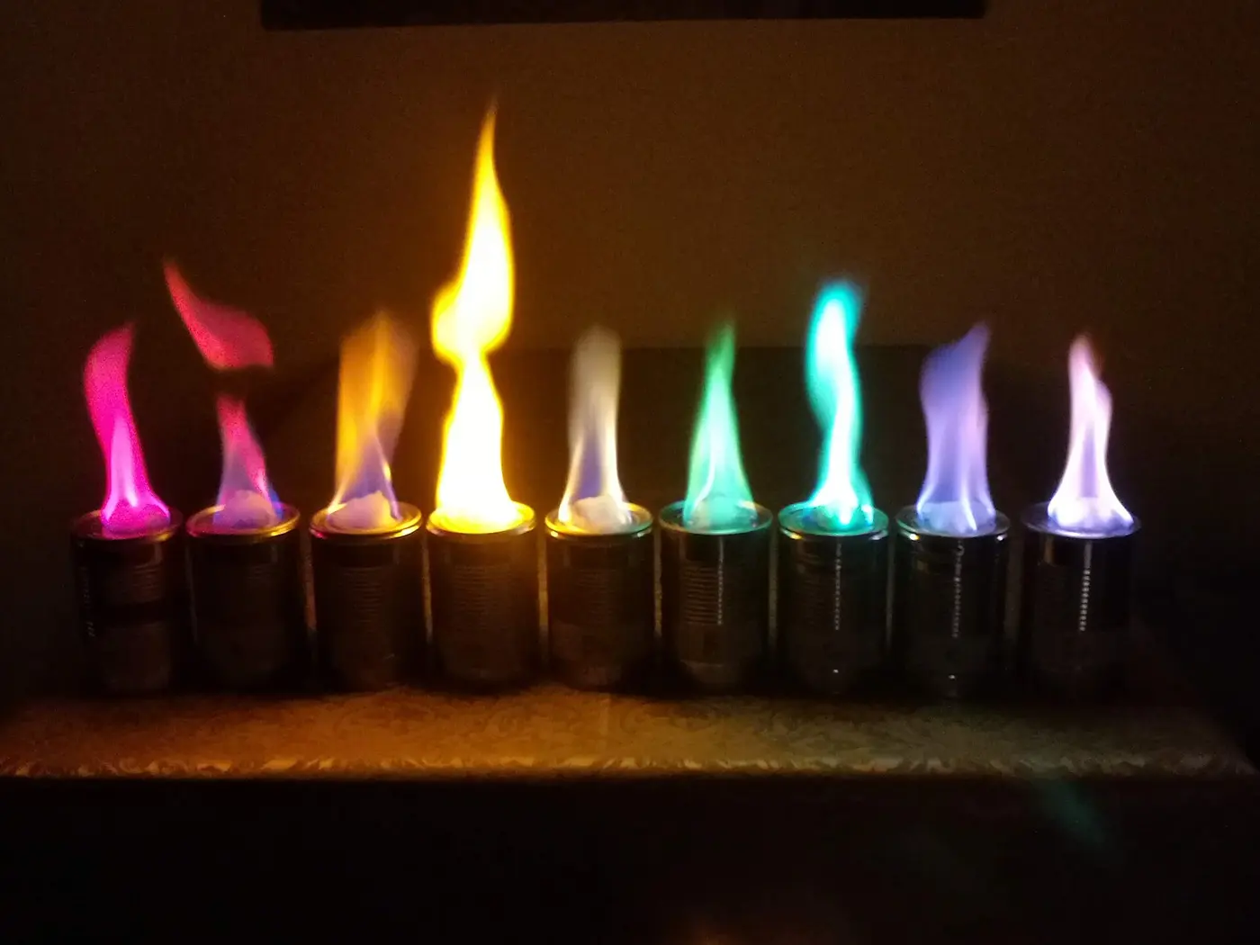 Nine flames in cans in a row, burning magenta, red, orange, yellow, whitish, green, blue-green, lilac and pink