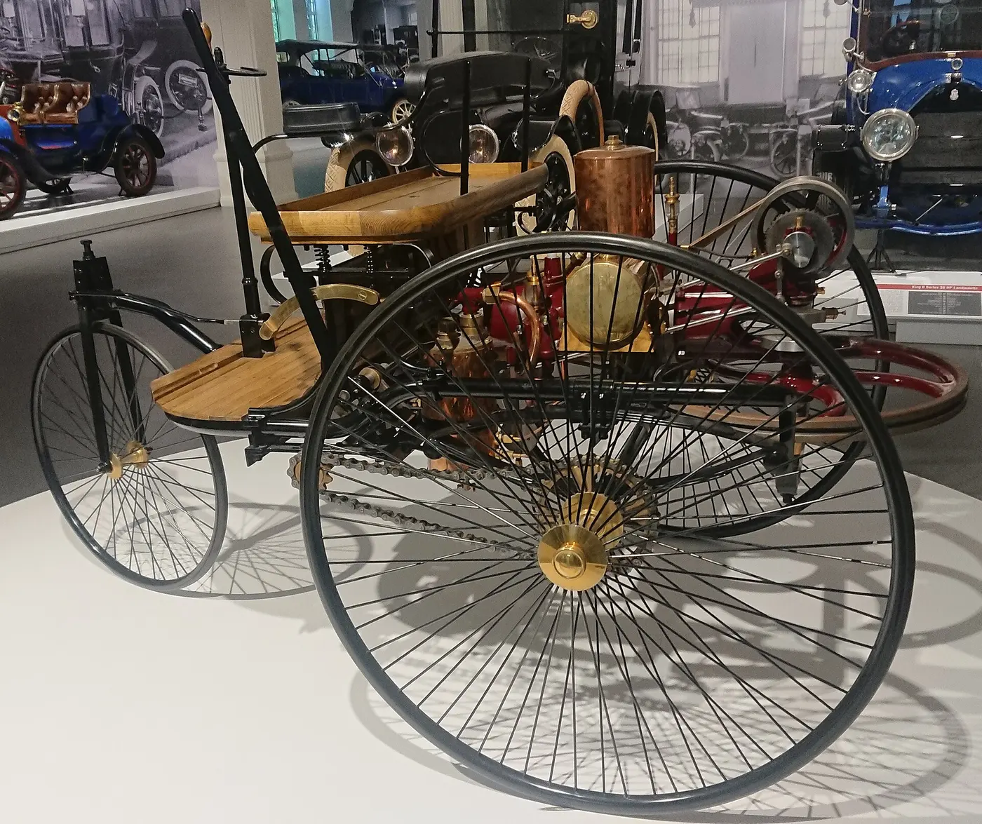 A vehicle with three large, thin-spoked wheels and a wooden seat, with a copper-colored engine mounted behind the seat