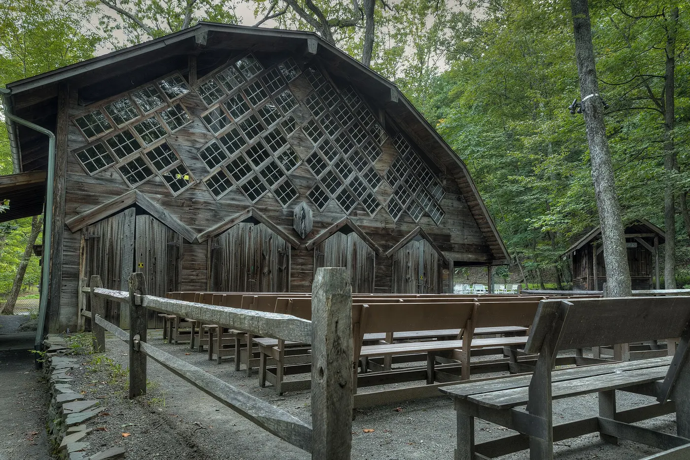 An old wooden hall with plank walls standing in the woods, its gable covered in diamond-shaped windows, with wooden benches in front