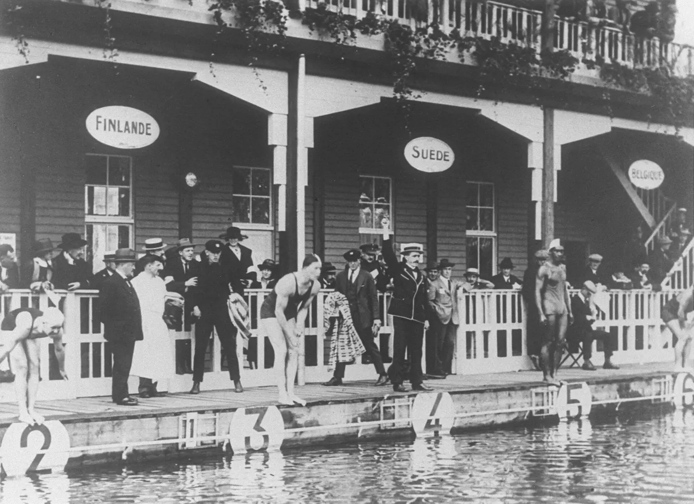An old black-and-white photo of swimmers lined up at the edge of an outdoor pool, waiting for the start