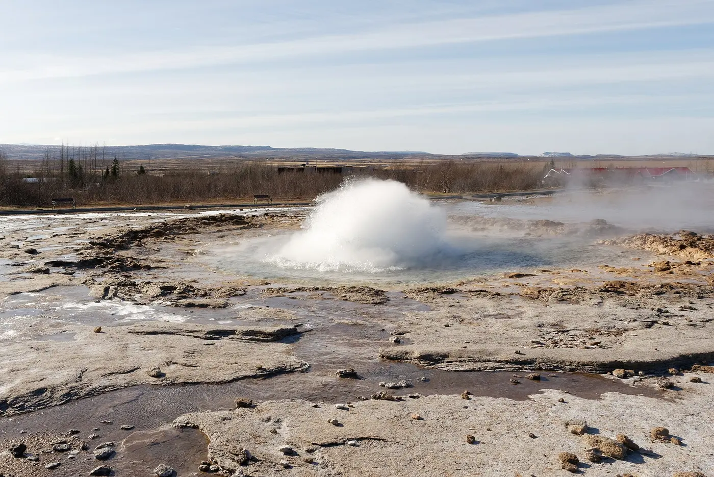 A geyser in the middle of steaming rocky ground, its water surface bulging into a dome and bursting white