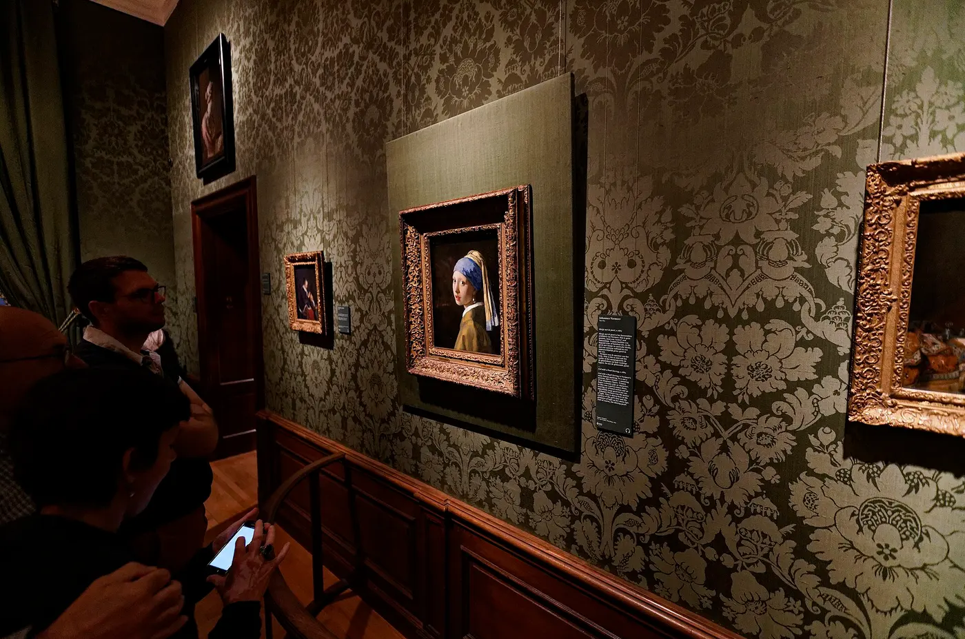 Visitors looking at Girl with a Pearl Earring hanging in a gallery with green patterned wallpaper