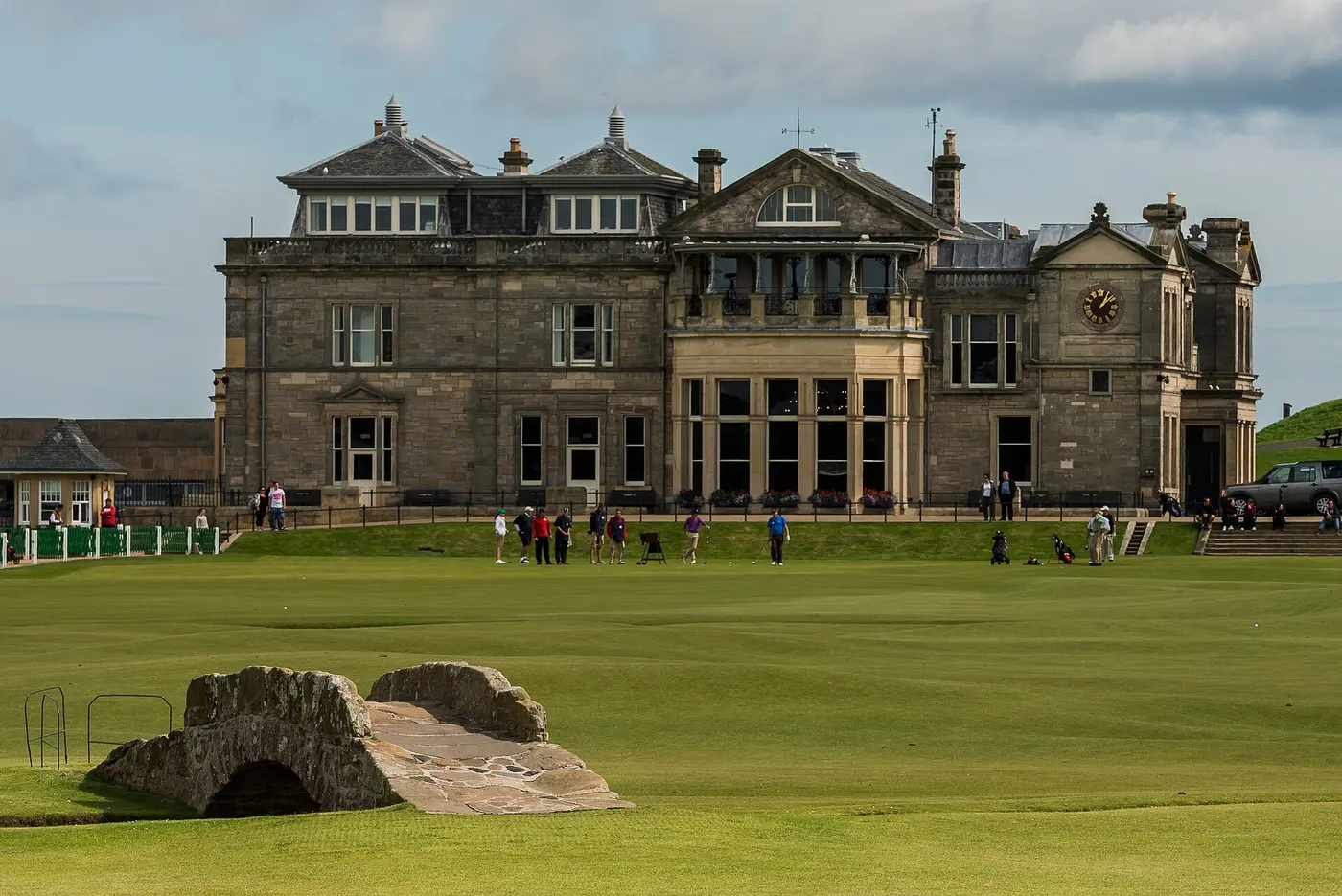 A small stone bridge over a grassy course, with a stone clubhouse behind it