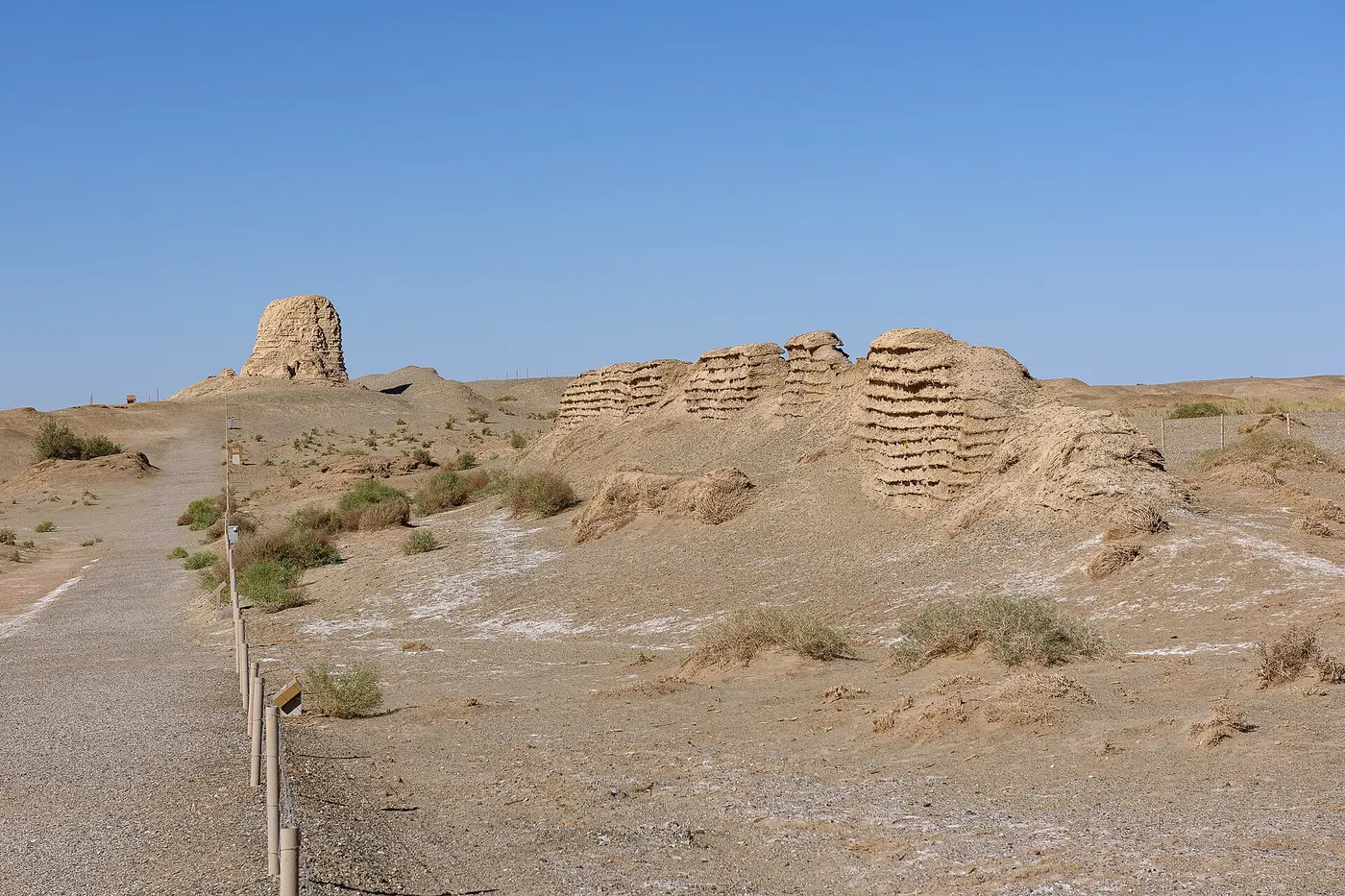 Low, earth-colored remains of a wall with visible horizontal layers, and the ruin of a square tower, stretching across dry wasteland under a blue sky