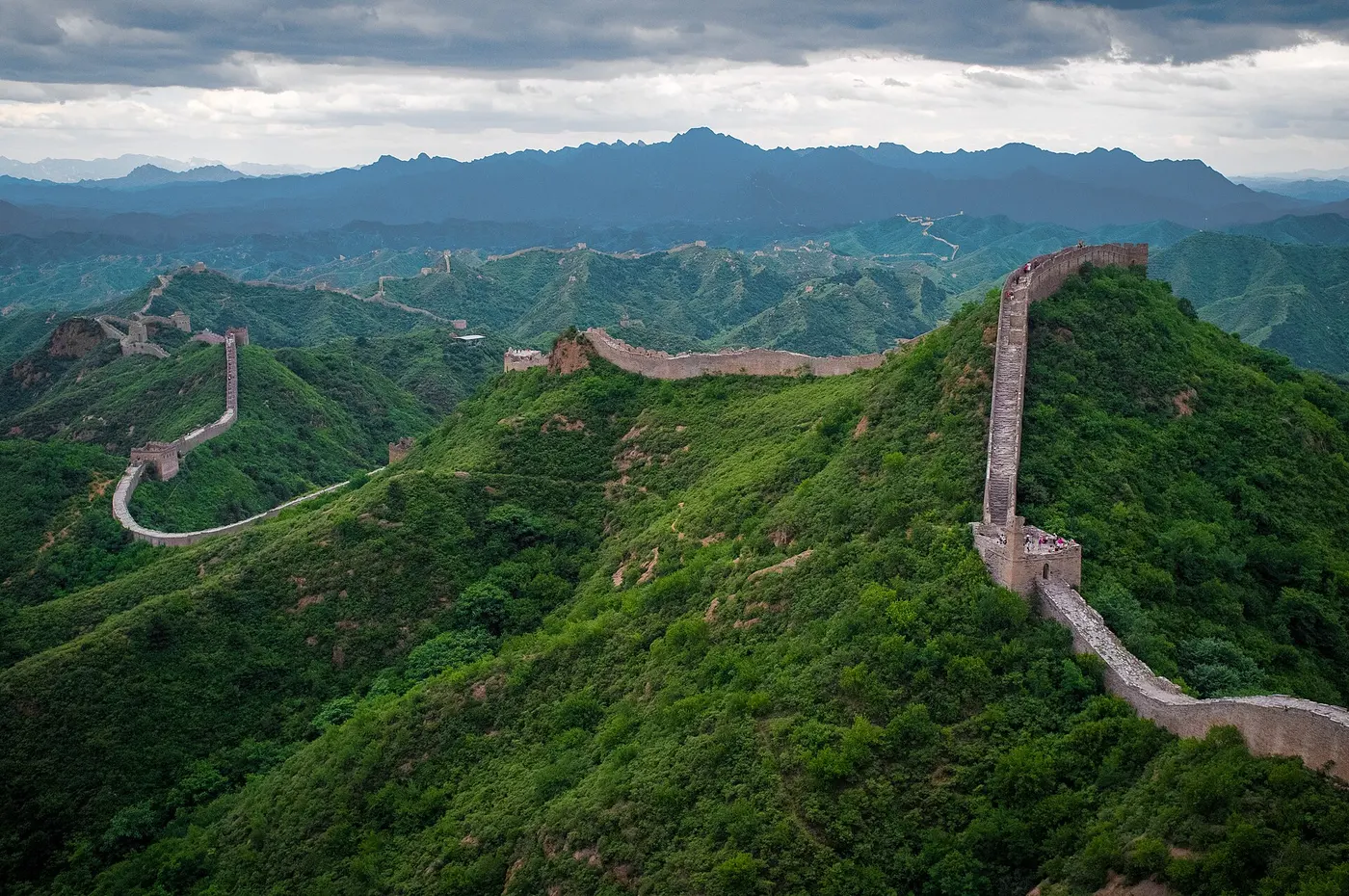A stone section of the Great Wall winding into the distance along a green, forested mountain ridge