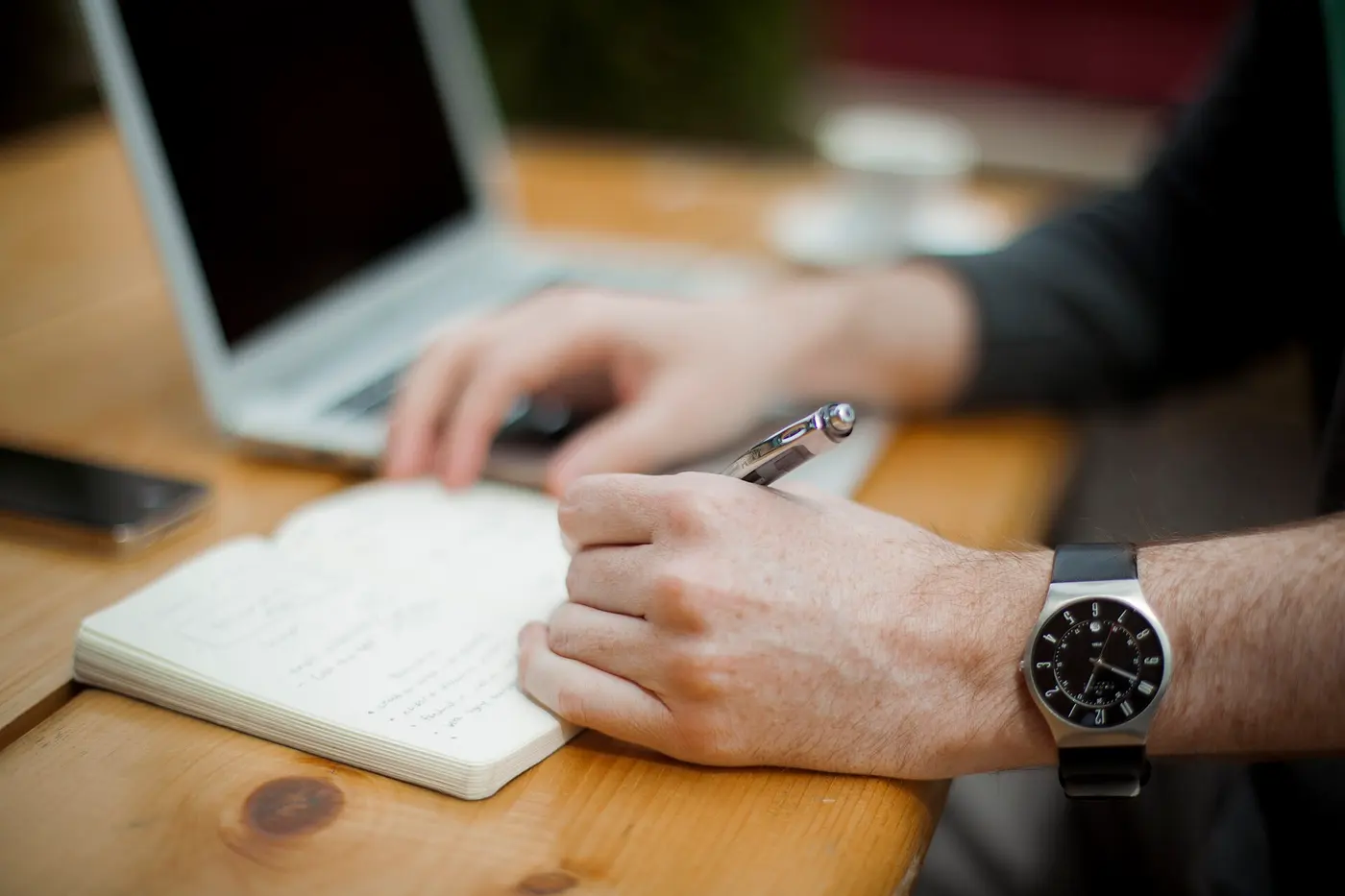 A close-up of a hand holding a pen in the left hand and writing in a notebook