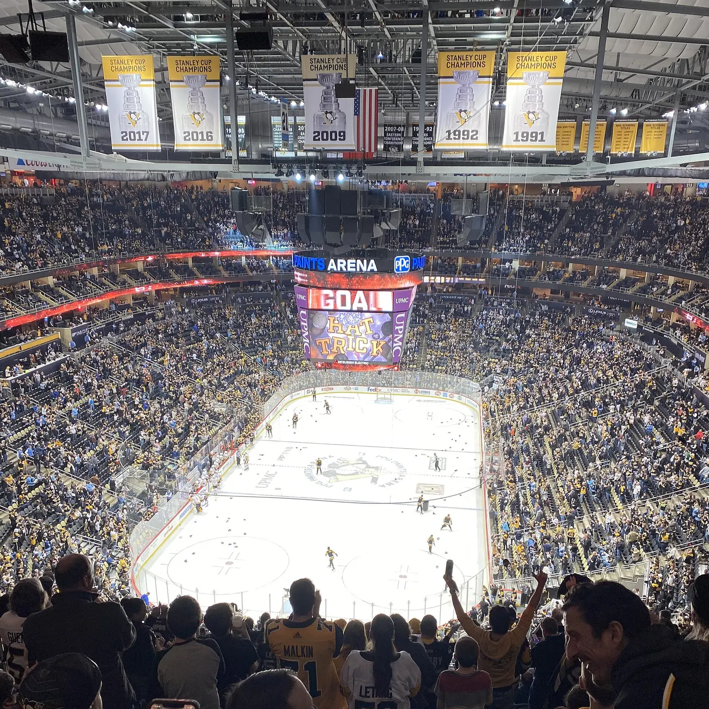 A packed ice hockey arena, with “HAT TRICK” on the big screen and dozens of dark hats scattered across the white ice