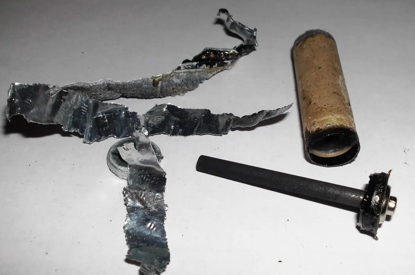 Parts of a disassembled battery: torn metal pieces, a cylinder and a black rod