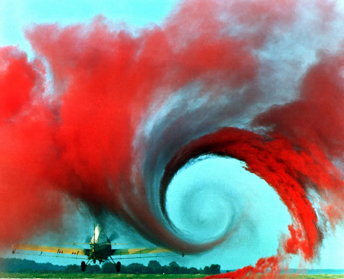 A small yellow biplane flying low through red smoke swirling into a large vortex