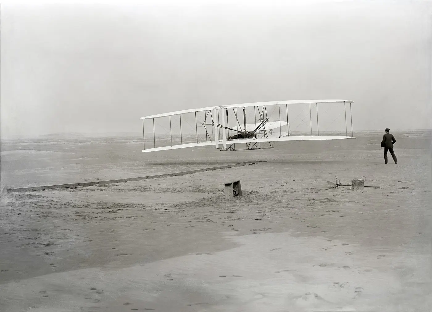 A black-and-white photo of a biplane flying low over sandy ground while a man looks on