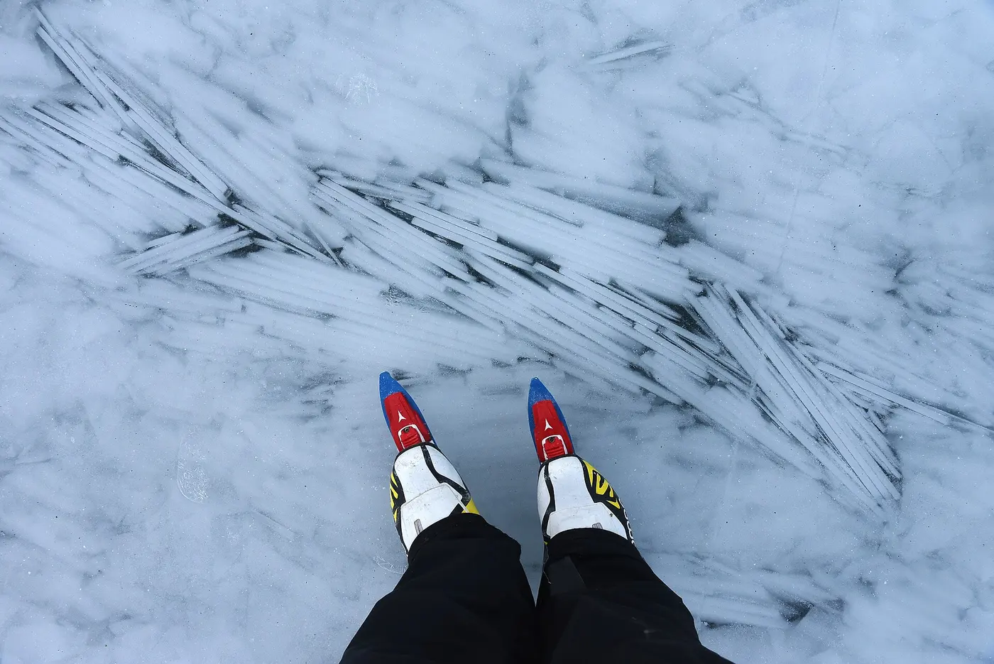 Two feet in skates with long, narrow blades stand on clear lake ice, with white plate-like patterns frozen inside