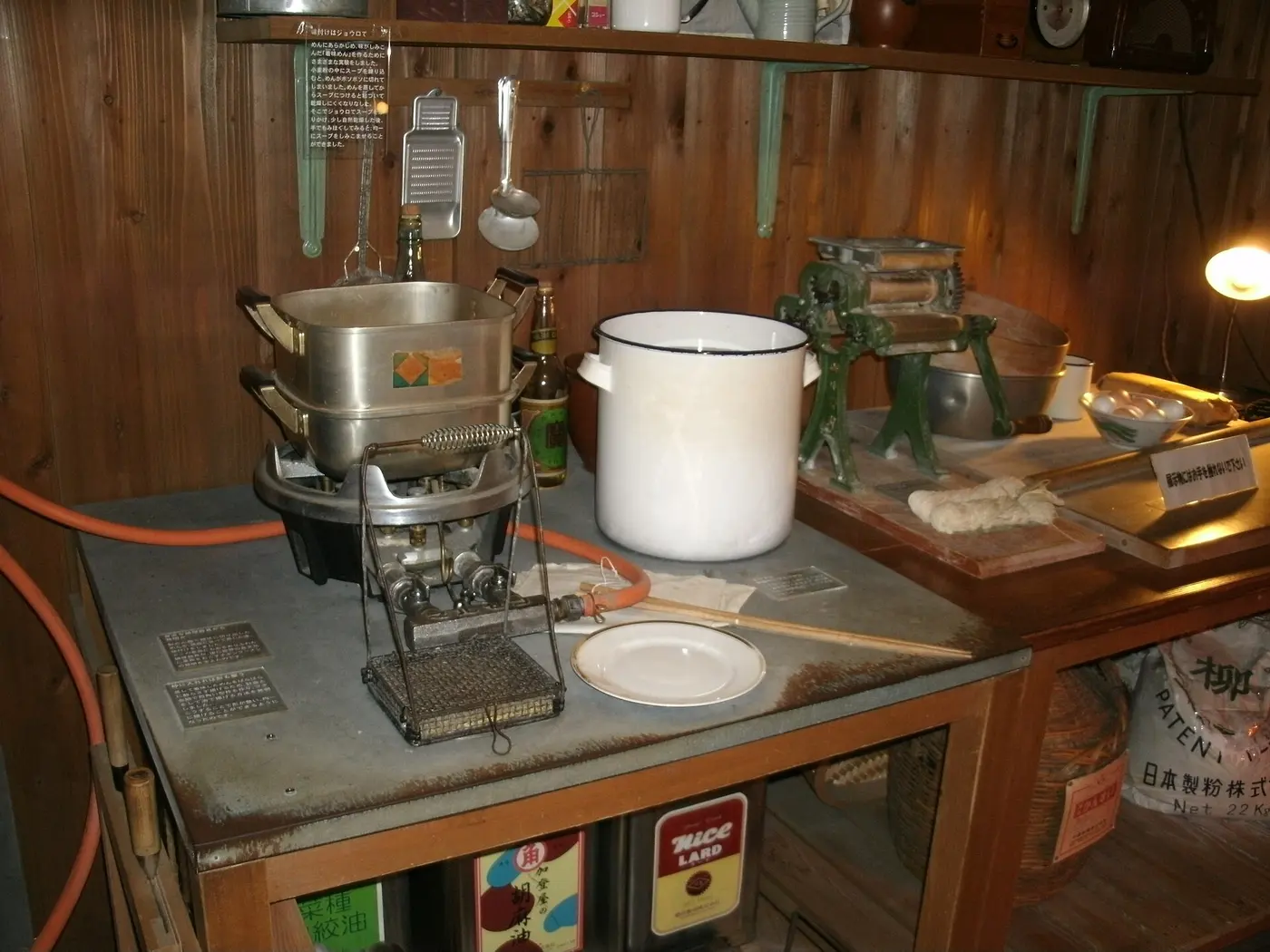 A gas burner, pots, a white enamel container and a noodle-making machine on a workbench in a wood-paneled room