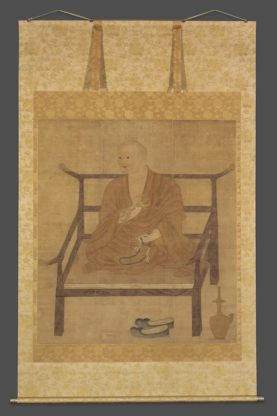 A hanging scroll in a gold-patterned mount, showing a monk in a brown robe sitting cross-legged on a wooden chair, holding a ritual implement and prayer beads
