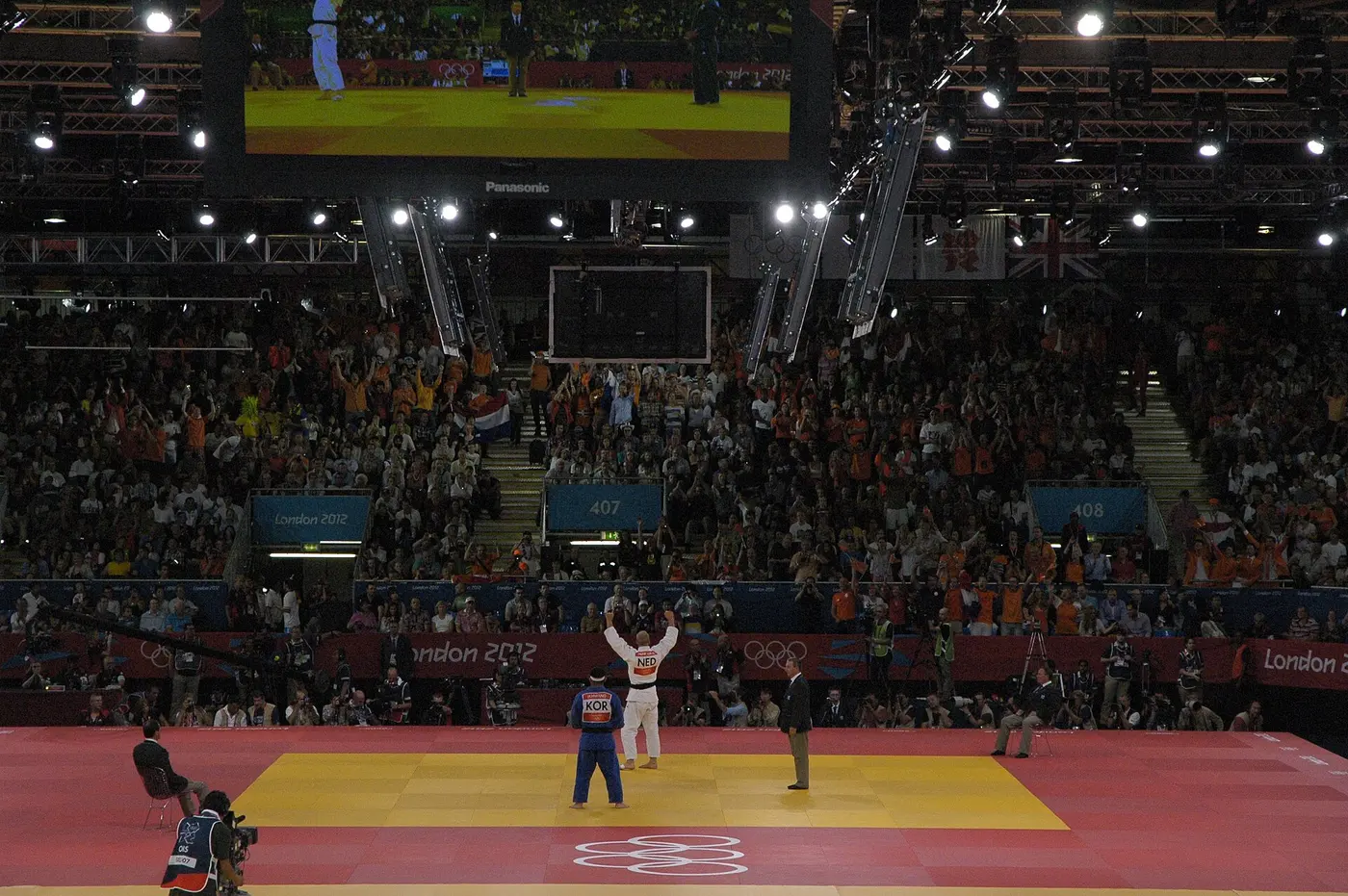 At the London Olympics judo venue, an athlete in a white uniform raises both arms, facing an athlete in a blue uniform