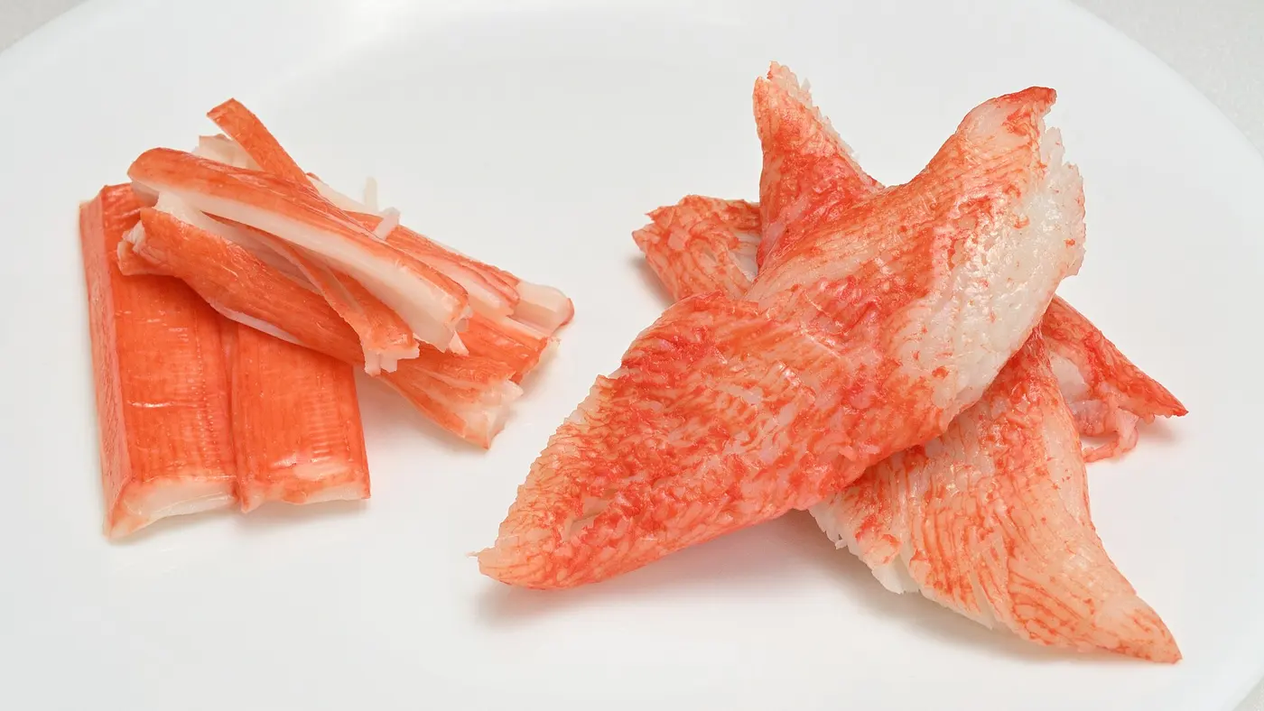 On a white plate, stick-shaped imitation crab on the left and chunks with a red surface and white interior on the right