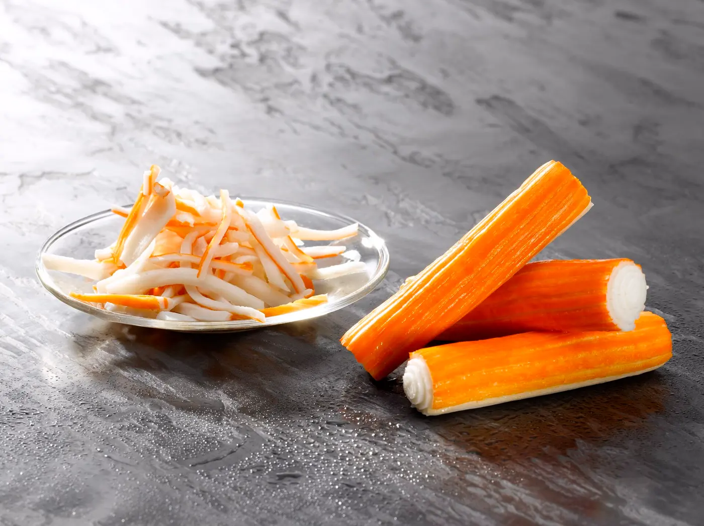 Three thick orange-striped imitation crab sticks on a gray surface, beside a small glass dish of shredded imitation crab