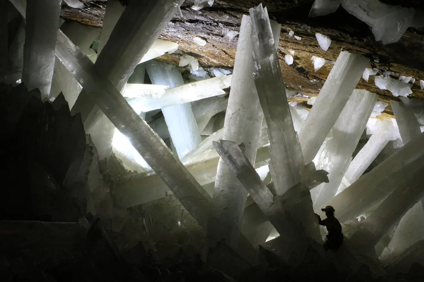 Huge white columnar crystals crisscrossing inside a cave, with a small human silhouette at their base