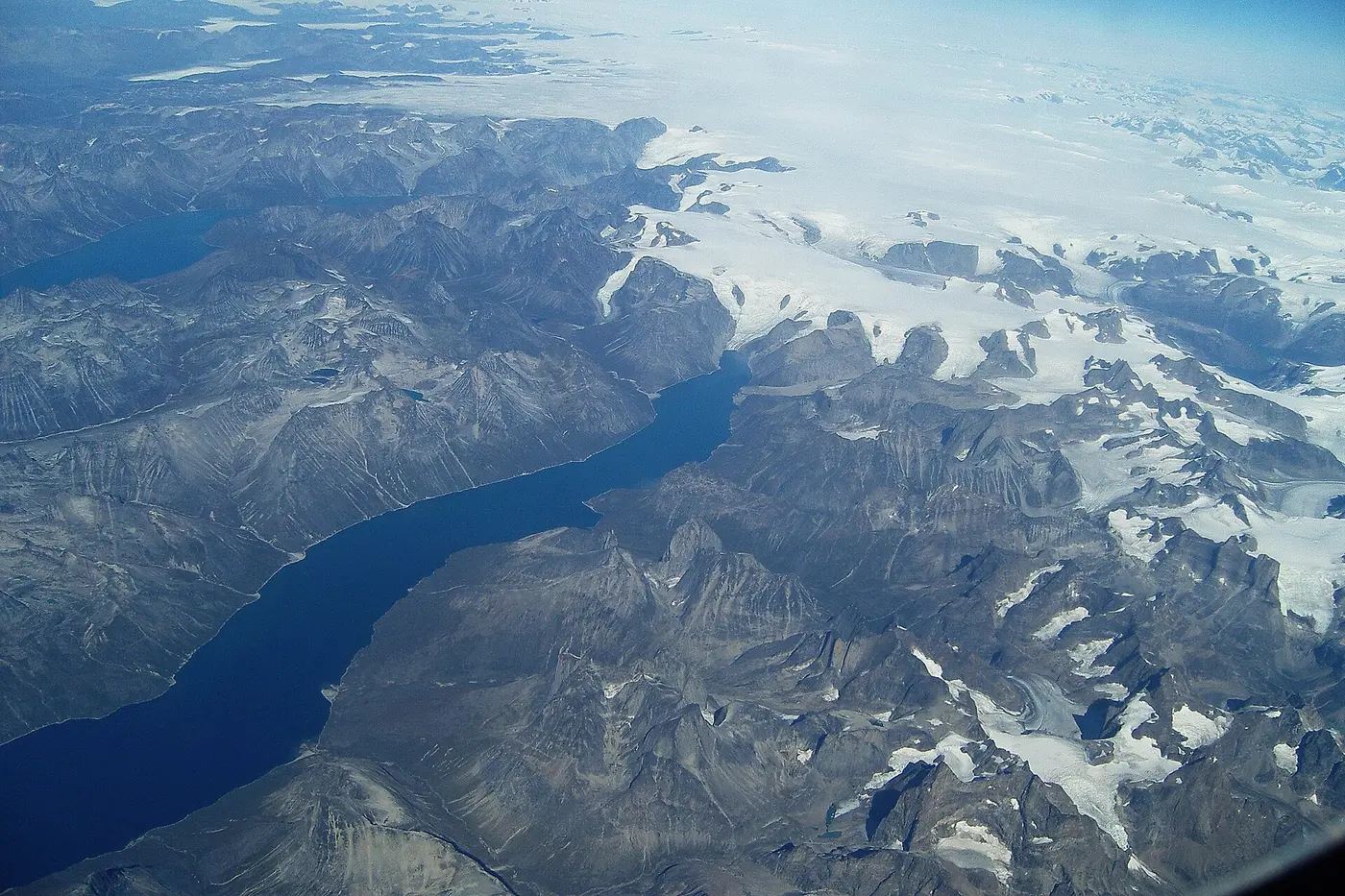 An aerial view of deep inlets and rugged rocky mountains, with a white expanse of ice beyond