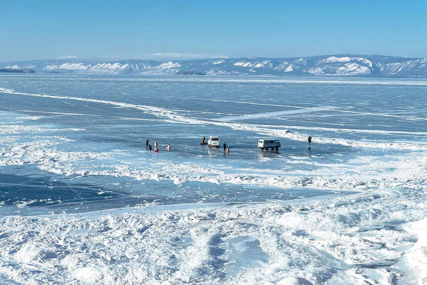 Cars and people dotted across a lake covered in blue ice, with snow-capped mountains behind